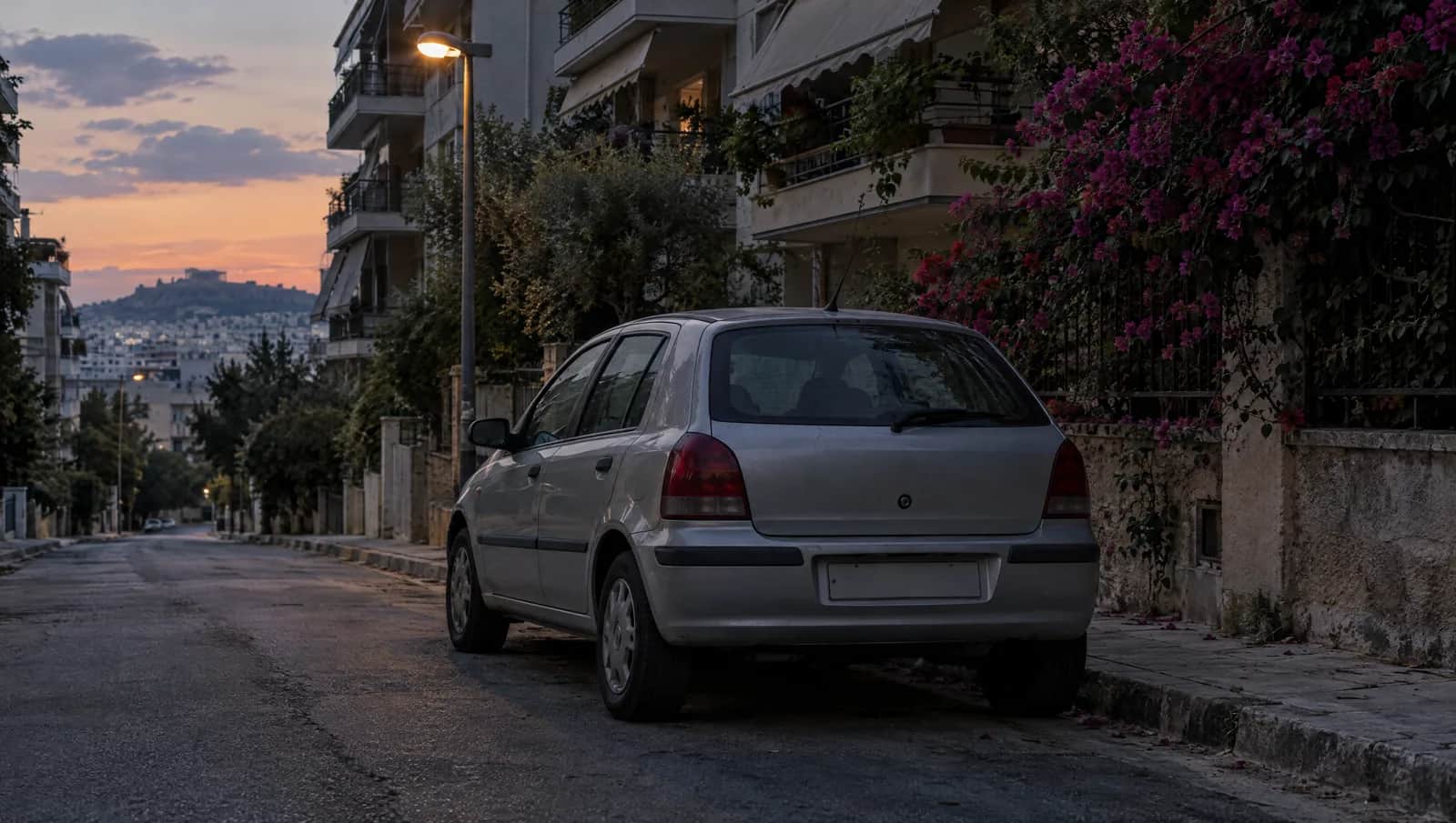 Ασημί μικρό αυτοκίνητο με κενή πινακίδα σταθμευμένο μόνο του σε ήσυχο δρόμο της Αθήνας το σούρουπο, με πολυκατοικίες και βουκαμβίλια πίσω του
