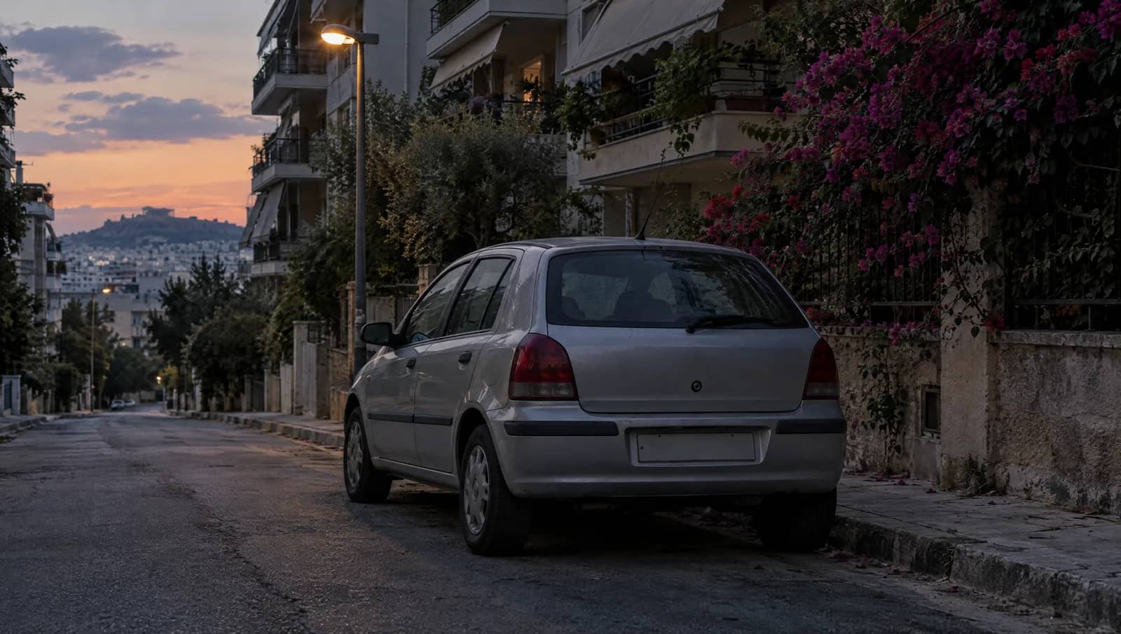 Ασημί μικρό αυτοκίνητο με κενή πινακίδα σταθμευμένο μόνο του σε ήσυχο δρόμο της Αθήνας το σούρουπο, με πολυκατοικίες και βουκαμβίλια πίσω του